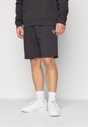 Mężczyzna ubrany w czarne szorty Reebok, czarną bluzę, białe skarpetki do kostki i białe trampki, stoi na jasnej podłodze na tle jednolitej ściany.