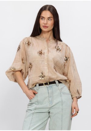 Vrouw met lang donker haar draagt een beige blouse met knopen en pailletten in bloemmotief en lichtblauwe high-waist jeans, poseert met één hand in haar zak.