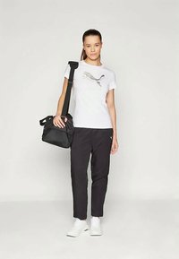 Joven mujer vistiendo una camiseta blanca de Puma, pantalones negros, zapatillas blancas, llevando un bolso negro en un fondo simple.