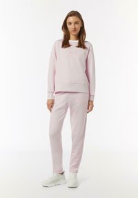 Rosa Sweatshirt mit besticktem Detail, kombiniert mit passenden rosa Jogginghosen. Weicher, gebürsteter Stoff, lockere Passform und gerippte Bündchen. Weiße Sneaker.