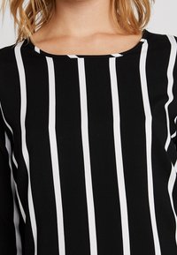 Kvinna i en svart långärmad topp med vertikala vita ränder och rundad halsringning, som visar övre bröst och axlar.