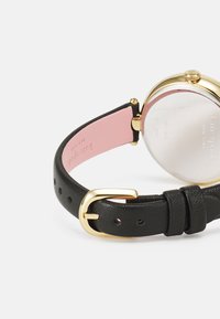 Svart läderrem för klocka med guldfärgad hårdvara, med en rosa läderfoder och en rund, reflexiv baksida med varumärkesgravyr.