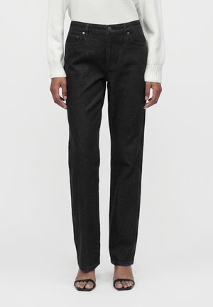 Kvinde iført sorte højtaljede jeans med lige ben og subtile glitrende detaljer, kombineret med en hvid top med lange ærmer og sorte sandaler med åben tå.