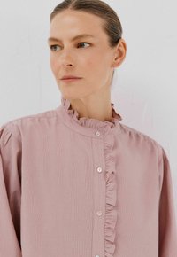 Donna con pelle chiara e capelli raccolti, indossa una blusa rosa a volant, guarda di lato contro uno sfondo semplice chiaro.