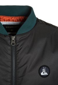 Černá bomber bunda s zeleným žebrovaným límcem, stříbrným zipem a kulatou černobílou nášivkou na levé části hrudi.