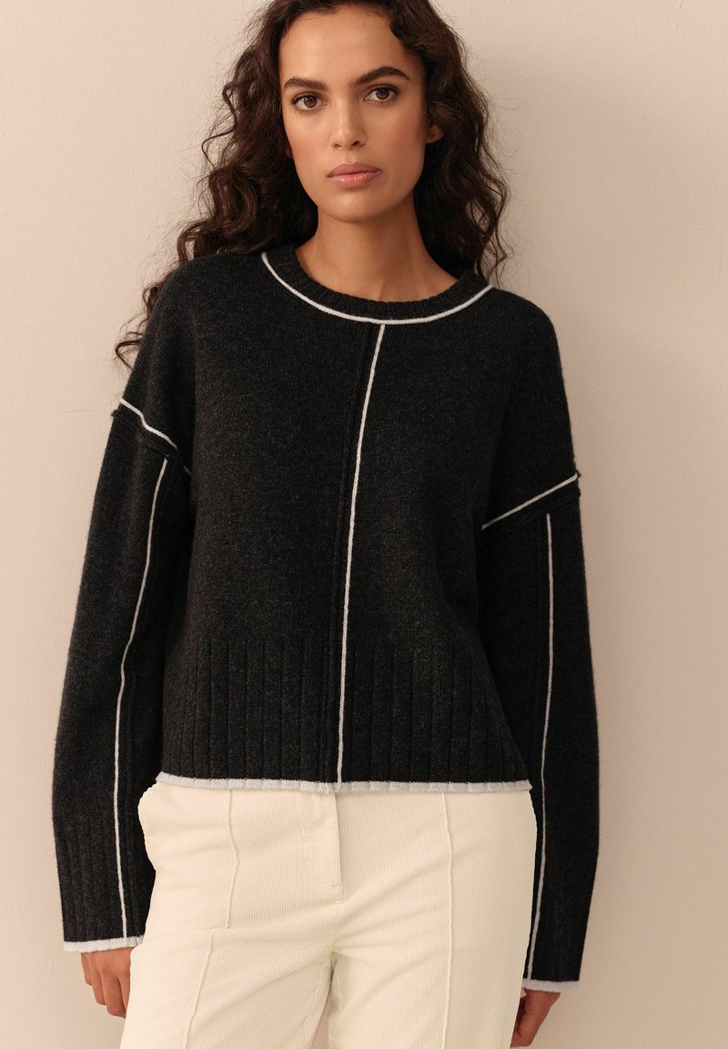 Maglione grigio scuro con cuciture bianche a contrasto, tessuto a costine sull'orlo e maniche larghe, abbinato a pantaloni chiari.