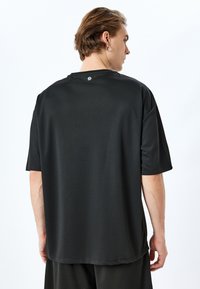 Camiseta deportiva negra con mangas cortas, textura suave, cuello redondeado y un pequeño logo en la parte posterior cerca del cuello.