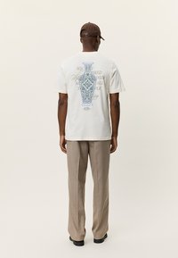 Λευκό βαμβακερό T-shirt με μπλε διακοσμητικό μοτίβο βάζου στην πλάτη, συνδυασμένο με μπεζ καρό παντελόνι και καφέ καπέλο.