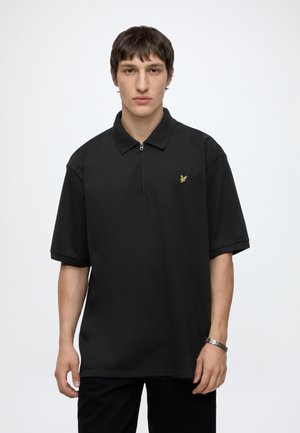 Ung mand med kort mørkt hår iført en sort poloshirt med korte ærmer og et lille gult fuglelogo på venstre bryst.
