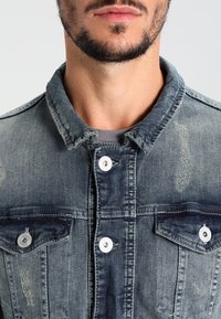 Džínová bunda v bledě modré barvě s límečkem, předními kapsami a bílými kovovými knoflíky. Obsahuje opotřebované oblasti a texturovanou tkaninu.