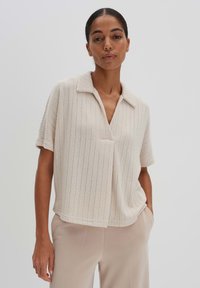 Kurzärmliges, kragenloses Oberteil in hellem Beige mit vertikalem Rippenmuster und lockerer Passform, kombiniert mit beigen Hosen.
