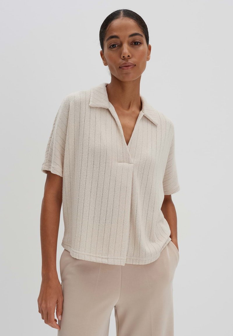 Kurzärmliges, kragenloses Oberteil in hellem Beige mit vertikalem Rippenmuster und lockerer Passform, kombiniert mit beigen Hosen.