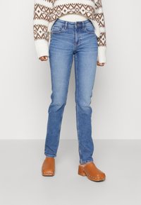 Vaqueros de mezclilla azul claro con un corte ajustado, que presentan un diseño clásico de cinco bolsillos y un desvanecimiento sutil. Combinados con zuecos de madera marrón.