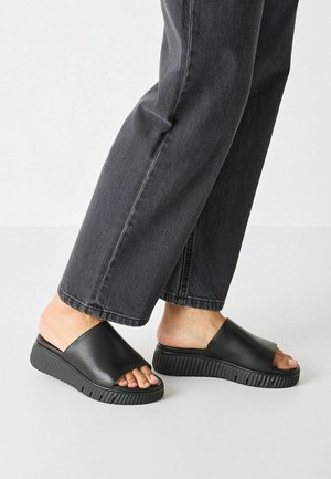 Pieds portant des sandales noires sans brides avec des semelles épaisses rainurées et un pantalon en jean gris foncé sur fond blanc.