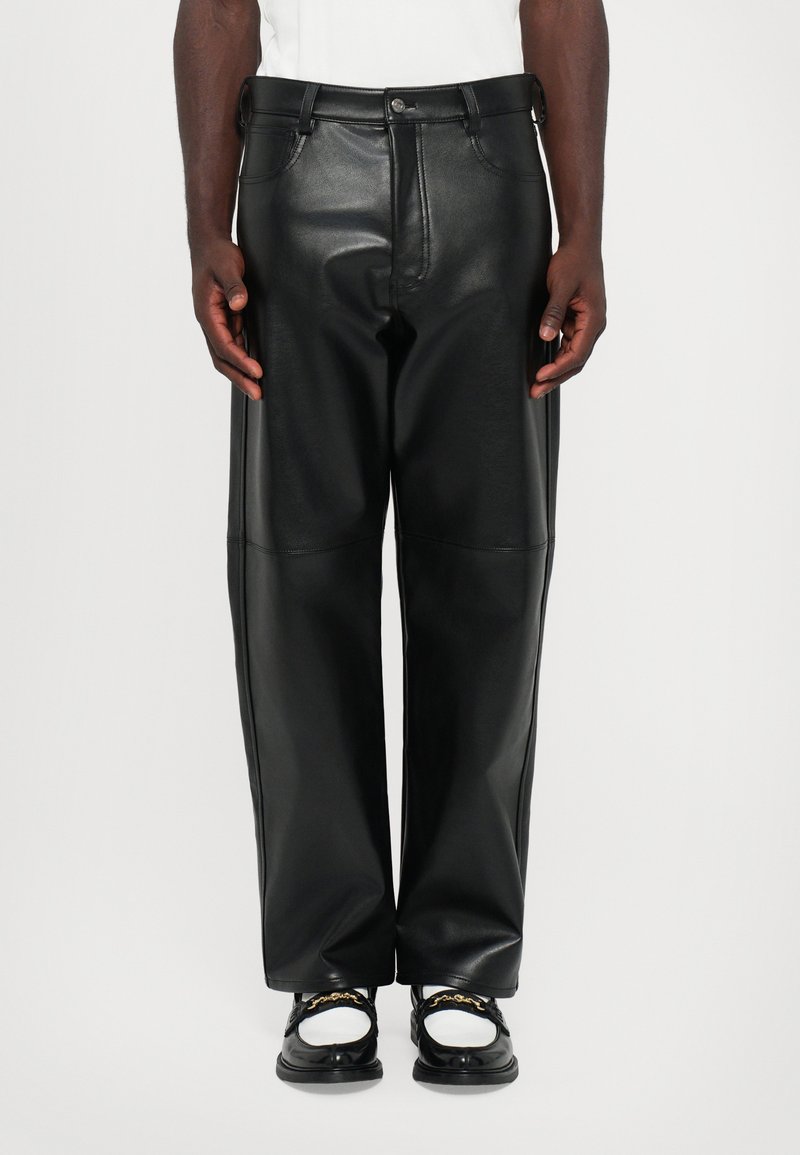 Homme portant un pantalon en cuir large noir et des mocassins noir et blanc avec des détails en chaîne dorée, se tenant devant un fond uni.