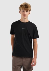 Tienerjongen in een zwart T-shirt met klein "BALLIN'" logo en een bruine broek, staande tegen een effen lichtgrijze achtergrond.