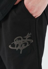 Black sweatpants made of soft fabric, featuring a prominent gray logo design on the left thigh and side pockets.