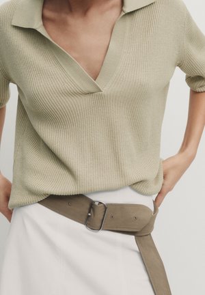 Nahaufnahme einer Person, die ein beiges, geripptes Strickoberteil mit kurzen Ärmeln und weitem Kragen trägt, kombiniert mit einem weißen Rock und einem breiten Taupe-Gürtel mit silberner Schnalle.