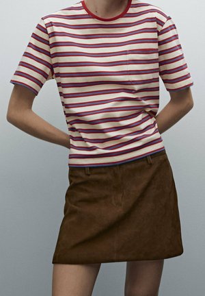 Personne portant un t-shirt à manches courtes rayé crème, rouge et bleu, rentré dans une mini-jupe en suède marron, les mains reposant derrière le dos.