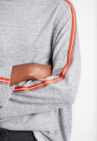 Personne à la peau foncée, les bras croisés, portant un pull gris avec des rayures orange et blanches le long des manches.