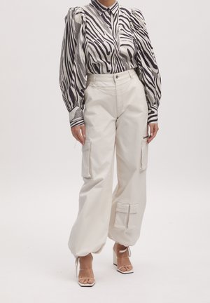 Femme portant un chemisier à manches bouffantes à motif zèbre, un pantalon cargo écru taille haute avec des poches, et des talons ouverts beige.