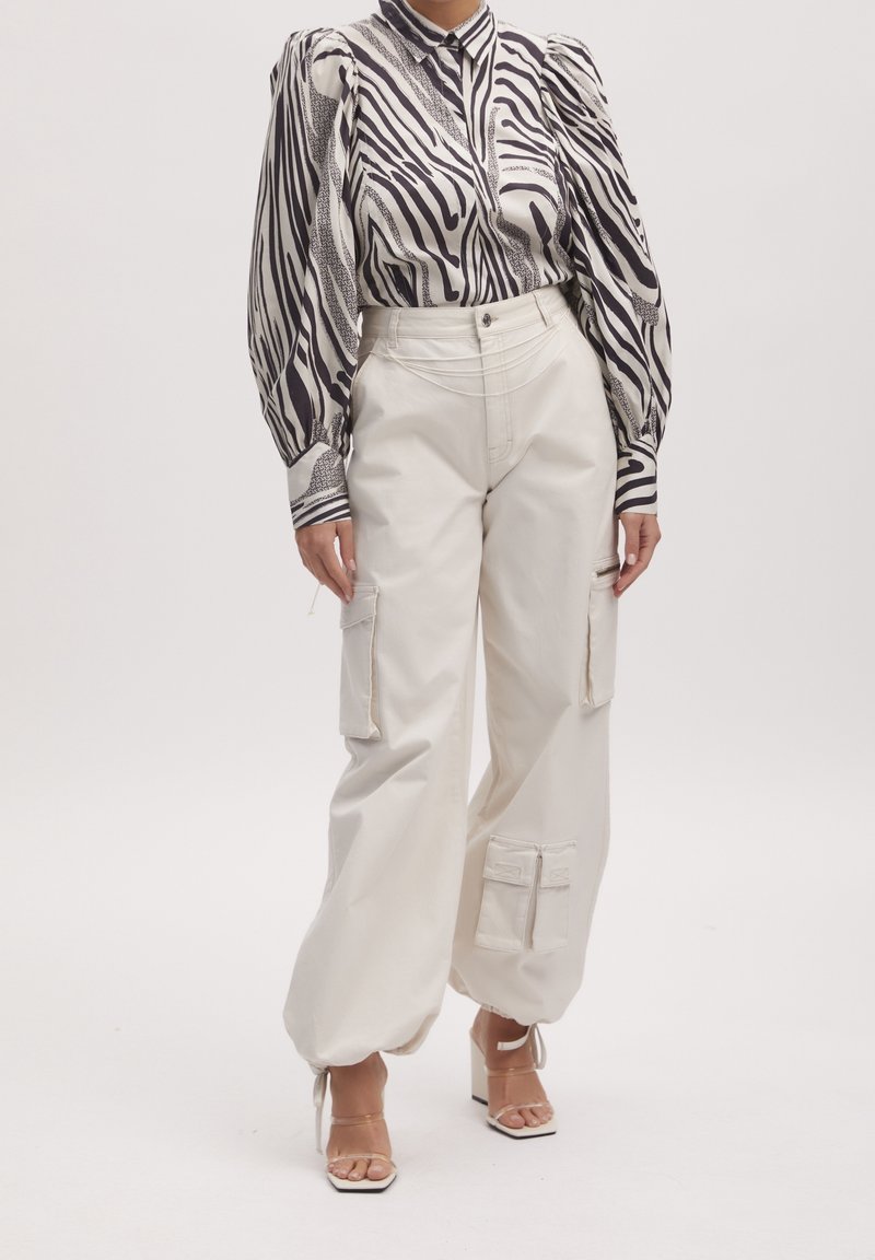 Femme portant un chemisier à manches bouffantes à motif zèbre, un pantalon cargo écru taille haute avec des poches, et des talons ouverts beige.