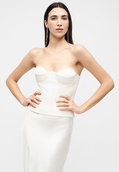 Γυναίκα με μακριά σκουρόχρωμα μαλλιά, φορώντας λευκό bustier τοπ χωρίς τιράντες και ταιριαστή φούστα, στέκεται με τα χέρια στη μέση μπροστά σε απλό φόντο.