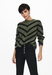 Gestreifter Pullover in Grün und Schwarz mit rundem Ausschnitt, langen Ärmeln und lockerer Passform, kombiniert mit hoch taillierten schwarzen Jeans mit Knopfdetails.