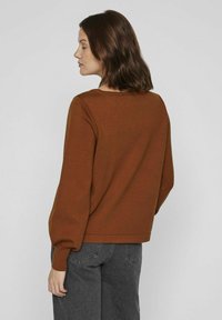 Pull-over marron avec un col côtelé et des manches longues légèrement bouffantes. Porté avec un jean taille haute gris. Texture lisse.