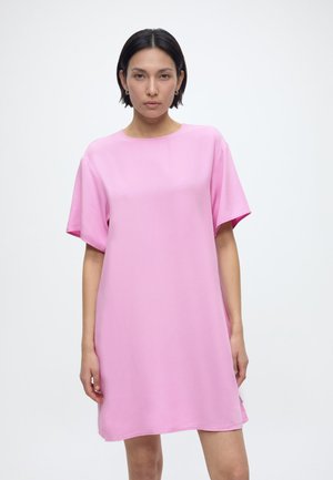 Kobieta o krótkich ciemnych włosach, ubrana w luźną różową sukienkę z krótkim rękawem, stoi na tle jednolitego jasnego tła.