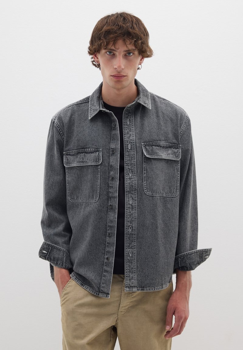 Chemise en denim gris avec un front boutonné, deux poches poitrine et des manches longues retroussées, assortie à un pantalon de couleur claire, sur fond blanc.