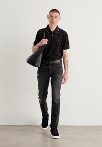 Schwarzes Polo-Shirt mit weißen Akzenten, dunkle Jeans und schwarze Sneaker. Das Modell trägt eine große schwarze Tasche. Der Hintergrund ist schlicht und minimalistisch.
