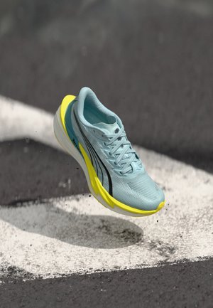 Chaussure de course bleu clair avec une tige en mesh respirant, présentant des rayures noires et une semelle jaune vif. En position aérienne, sur une surface de piste.