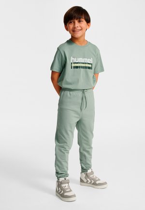 Garçon souriant debout les mains dans le dos, portant un t-shirt et un pantalon de sport hummel verts, baskets montantes blanches et grises.