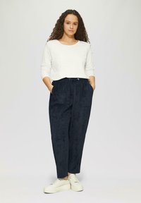 Witte lange mouwen top, marineblauwe sportbroek met zakken en trekkoord, gecombineerd met witte chunky sneakers, tegen een neutrale achtergrond.