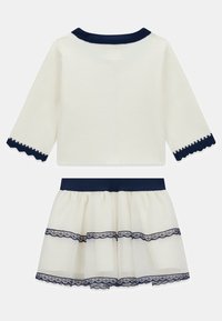 Ensemble deux pièces : haut à manches longues crème avec accents bleu marine, poignets festonnés, et jupe assortie à volants avec bordure en dentelle. Tissu doux, design féminin.