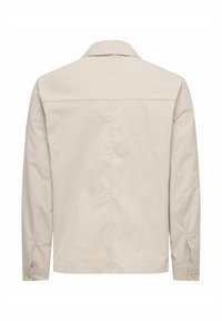 Veste beige clair avec un col, manches longues, poignets à boutons et une texture lisse. Le dos présente une empiècement et des détails de couture soignés.