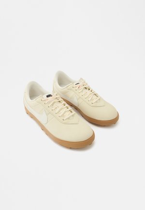 Valged beežid Nike tossud, millel on sile nahast pealmine osa, paelaga kinnituv ja kummist jope. Omaduseks on kontrastse valge Swooshi logo.