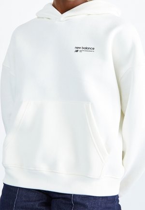 Osoba ubrana w białą bluzę New Balance z kapturem i kieszenią z przodu oraz dżinsy, widoczne logo na piersi.
