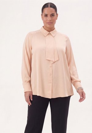 Femme aux cheveux foncés attachés en un chignon élégant, portant un chemisier rose pâle à manches longues avec un détail de nœud à l'avant et un pantalon noir sur un fond clair.