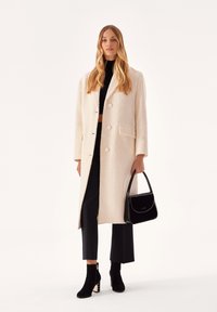 Cappotto lungo di colore crema con una texture morbida e grandi bottoni, abbinato a pantaloni neri e stivaletti alla caviglia con tacco a blocco e zip.