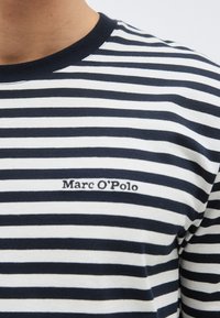 Κοντινό πλάνο ενός ατόμου που φοράει μπλε ναυτικό και λευκή ριγέ μπλούζα με γιακά σε μπλε ναυτικό και το κέντημα "Marc O'Polo" στο στήθος.