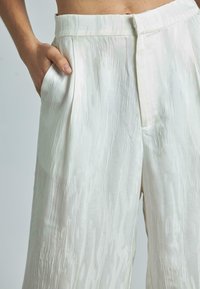 Pantalons blancs à la coupe large, texturés et légèrement brillants, dotés d'une taille plate et de poches sur les côtés. Un motif de lignes verticales ajoute un intérêt visuel.