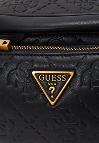 Černá kožená taška Guess se zlatým trojúhelníkovým logem, embosovaným vzorem a zlatým jezdcem zipu s výřezem ve tvaru trojúhelníku.