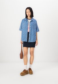 Cămașă din denim albastru cu mânecă scurtă și un model texturat, cu două buzunare frontale, purtată alături de o fustă mini bleumarin și pantofi cu platformă maro.