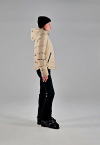 Person wearing beige puffer jacket, black pants, black beanie, and black shoes stands in profile against a plain gray background.