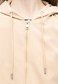 Veste beige à capuche avec une fermeture éclair à l'avant et des cordons de serrage, portée par-dessus un vêtement intérieur noir.