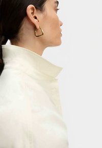 Gold hoop earring with a triangular shape worn on the ear, paired with a high-collared white shirt made of a smooth, light fabric.