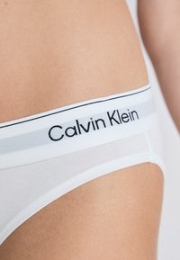 Slip di cotone bianco con una vita stretta. La vita presenta il marchio "Calvin Klein" in nero, con una striscia grigia sopra. Texture liscia.