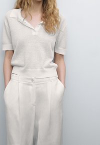 Neulepolo vaaleanbeige väreissä, jossa on kolmonen napitus ja lyhyet hihat, yhdistettynä korkeavyötäröisiin, vaaleanbeigeihin housuihin, joissa on laskokset.